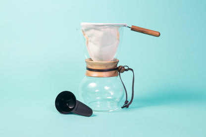 Glass server with a round base, acacia wood collar with leather tie, cloth filter and handle on top and a black plastic scoop.