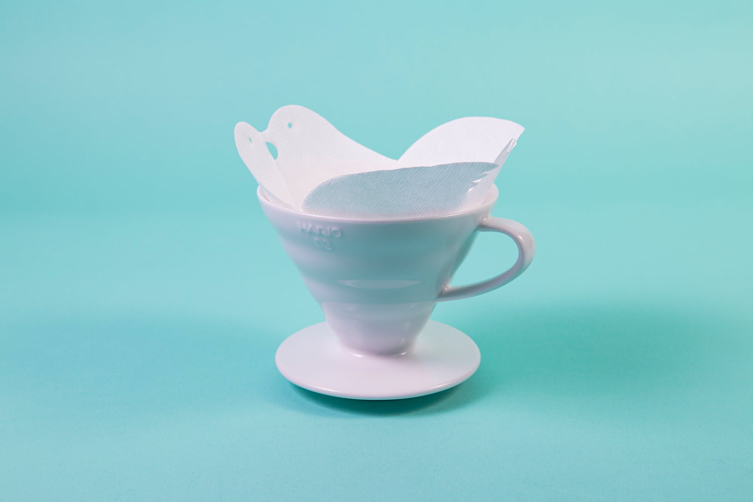 White cone paper filter, shaped into two birds kissing at the top, seated in a white ceramic cone-shaped dripper and set against a blue background.