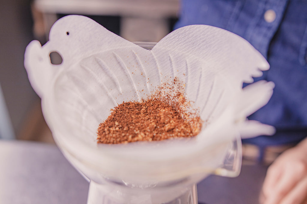 Close up of coffee grounds inside a white cone filter, shaped into two birds kissing at the top, and seated in a clear glass cone-shaped dripper. A person in blue button shirt in the background.