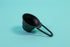 Matte black beehive shaped measuring spoon with bezier shaped open air handle on a blue background.
