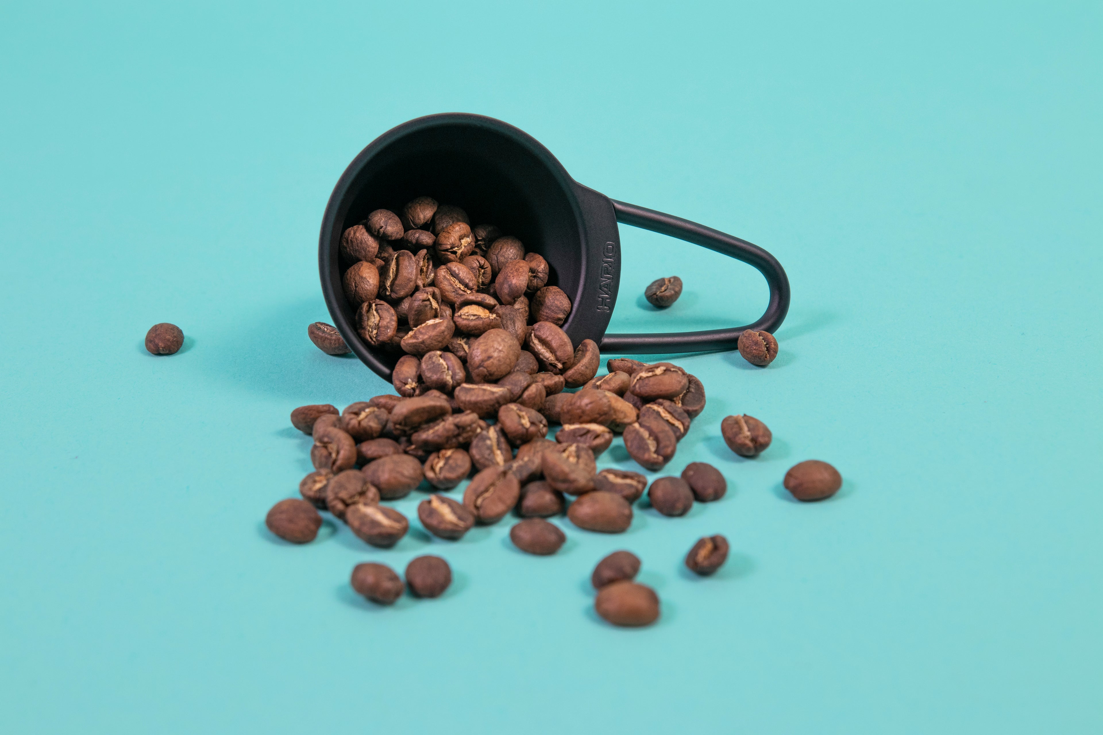 Matte black beehive shaped measuring spoon with bezier shaped open air handle with coffee beans spilling out on a blue background.