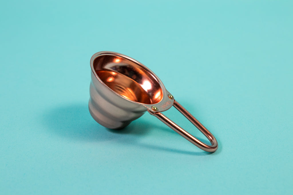 Copper beehive shaped measuring spoon with bezier shaped open air handle on a blue background.