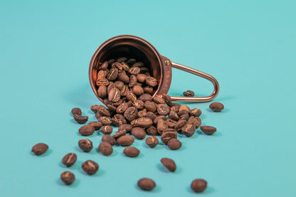 Copper beehive shaped measuring spoon with bezier shaped open air handle with coffee beans spilling out on a blue background.