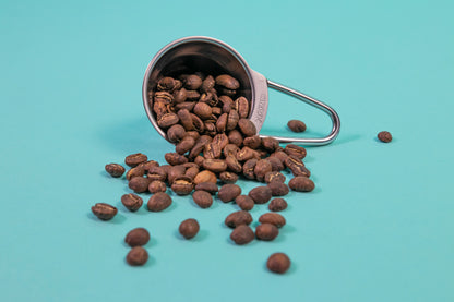 Stainless steel beehive shaped measuring spoon with bezier shaped open air handle with coffee beans spilling out on a blue background.