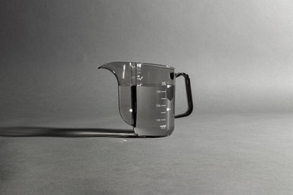 Transparent black plastic cup with handle and gooseneck style pour spout with water inside on a black and white background..