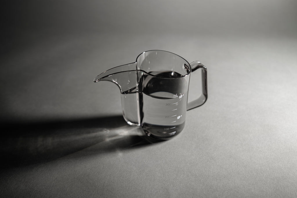 Transparent black plastic cup with handle and gooseneck style pour spout with water inside on a black and white background.