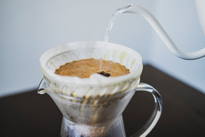 Close up of white gooseneck spout pouring water into brewing coffee inside a clear cone-shaped paper filter seated inside a tall, tapered glass server with fluted spout and full glass handle on a dark wood table.