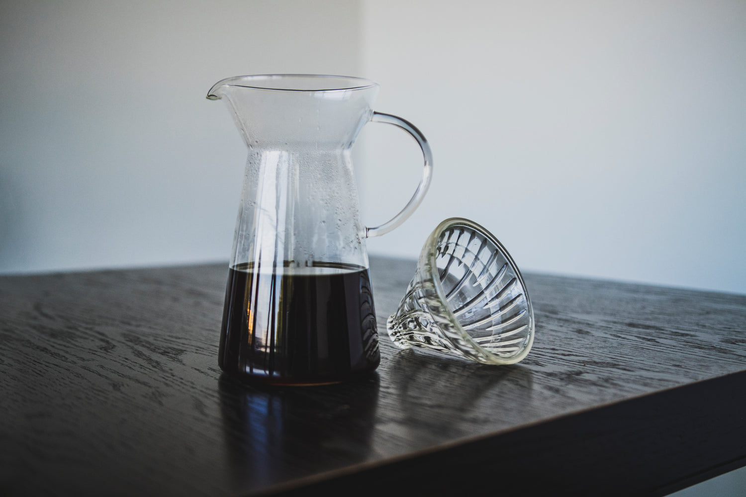 Tapered glass coffee server with fluted spout and all glass handle with coffee inside and a clear glass dripper with clear silicone ring seated beside on a dark wood table.