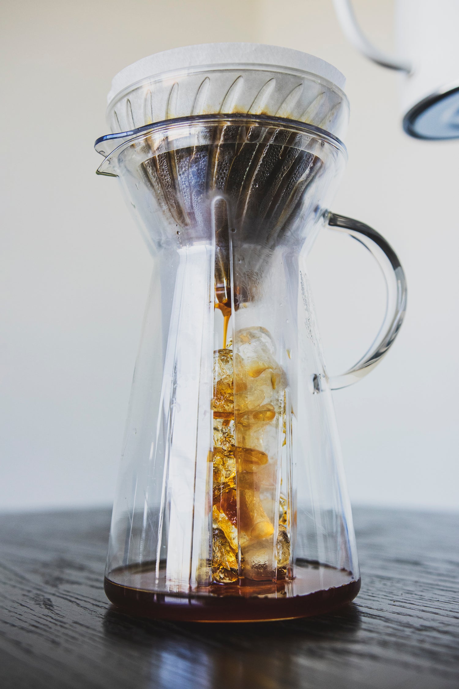 Coffee brewing over ice in a tall, tapered glass server with fluted spout and all glass handle with a white paper cone shaped filter in a glass cone shaped dripper with brewed coffee inside.