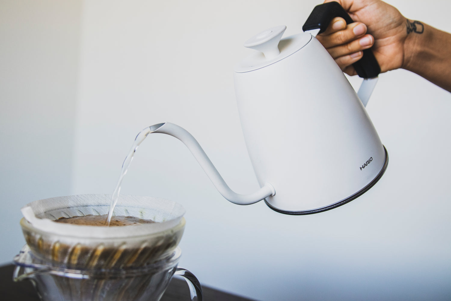 Hand pouring a matte white metal gooseneck kettle with white lid knob and black rubber handle cover into a glass cone-shaped filter with blooming coffee and seated in a glass pitcher.