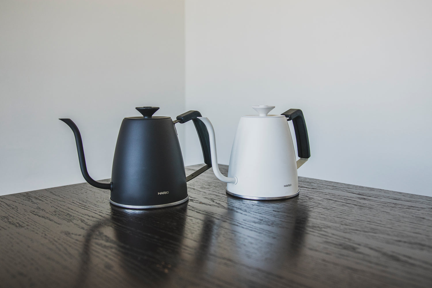 A matte whitemetal gooseneck kettle with white lid knob and black rubber handle cover and a matte black metal gooseneck kettle with black lid knob and rubber handle cover set on a black wood table with white background.