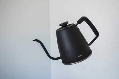 Matte black metal gooseneck kettle with black lid knob and rubber handle cove floating in the air in a titled pouring position.