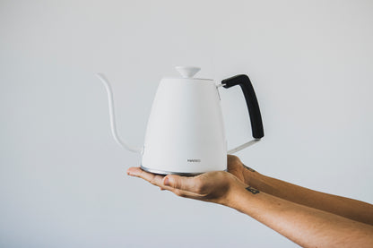Two hands holding a matte white metal gooseneck kettle with white lid knob and black rubber handle cover.