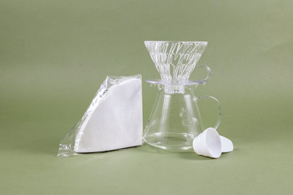 Glass coffee dripper with clear glass plastic base sitting on a tapered glass coffee picther with full glass handle and white paper filters in clear plastic packaging and white plastic coffee measuring spoon set against a green background.
