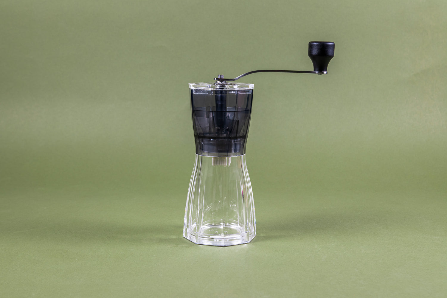 Clear plastic resin coffee hand grinder with hour-glass, octogonally shaped body, black clear plastic hopper, clear plastic lid, stainless steal handle, and black plastic knob set against a green background.