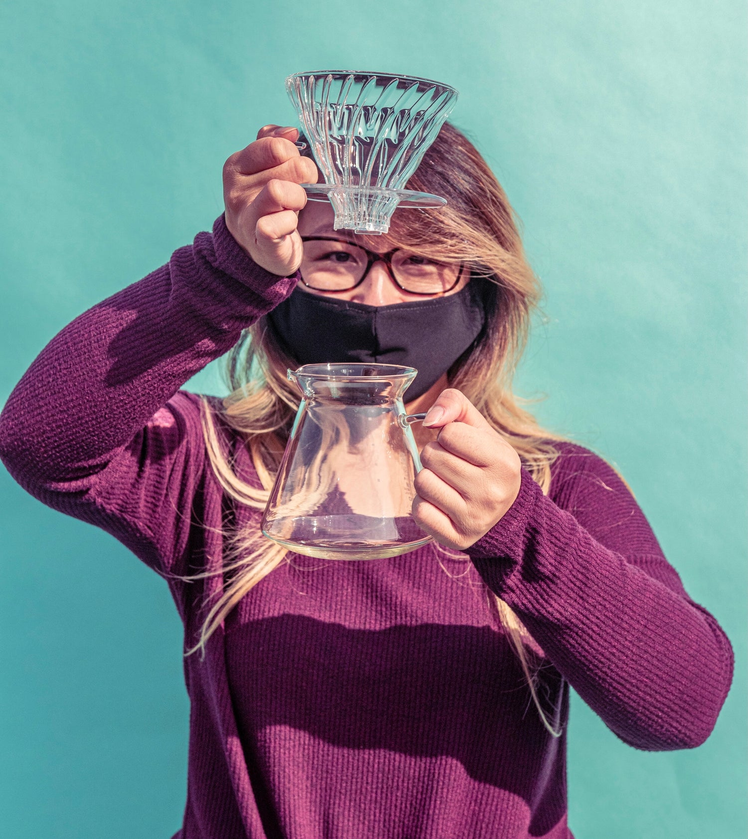 Woman in purple sweater and black face mask holding clear glass dripper with clear glass plastic base above tapered glass coffee pitcher with full glass handle and flare spout against a blue background.