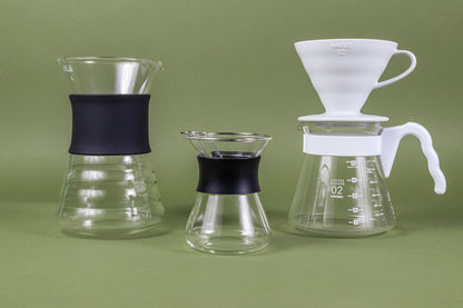 Tall hour glass shaped glass coffee server with beehive shaped bottom and black silicone band on left, hourglass shaped glass server with stainless steel wire dripper and black silicone rubber band in center, and white tapered glass coffee pitcher with white plastic handle and white ceramic cone-shaped coffee dripper seated on top on the right, and all set against a green background.