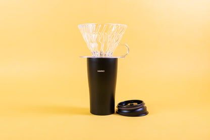 Clear glass cone-shaped coffee dripper with clear plastic round base and handle sitting on top of a textured black coffee tumbler with Hario logo and black lid resting upside down beside the tumbler and all set against a yellow background.