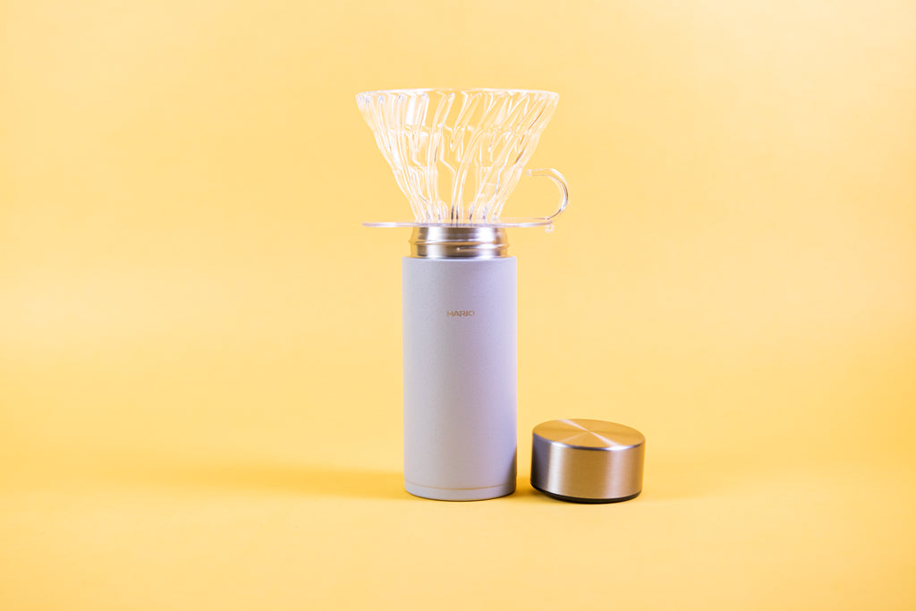 Slim cylindrical textured grey insulated cup with clear glass dripper with clear plastic base and handle seated on top and stainless steel lid resting beside. Set against a yellow background.