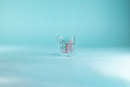 Glass measuring cup with small spout and measurement markings in red and black on the inside and outside of the glass set against a blue background.