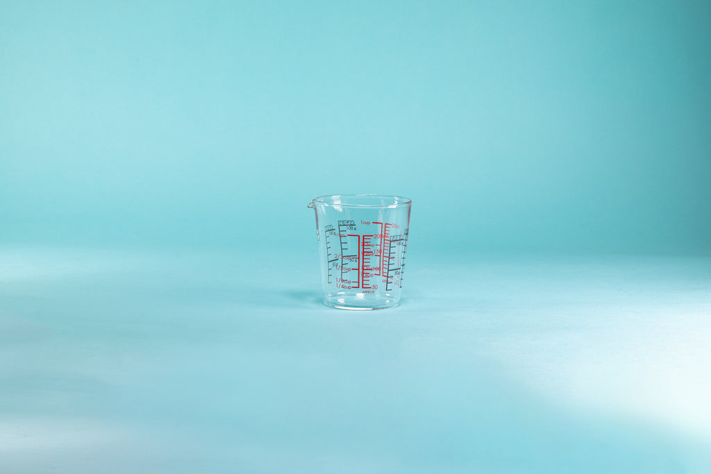 Glass measuring cup with small spout and measurement markings in red and black on the inside and outside of the glass set against a blue background.
