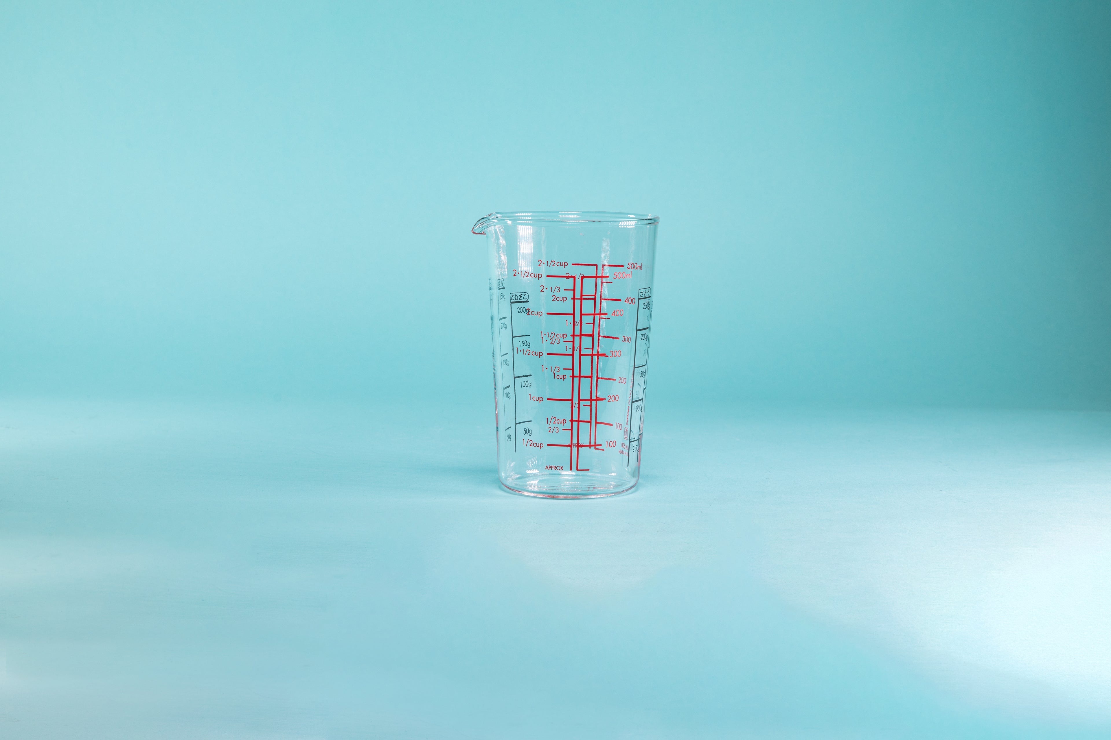 Tall cylindrical measuring cup with small spout and measurement markings in red and black on the inside and outside of the glass set against a blue background.