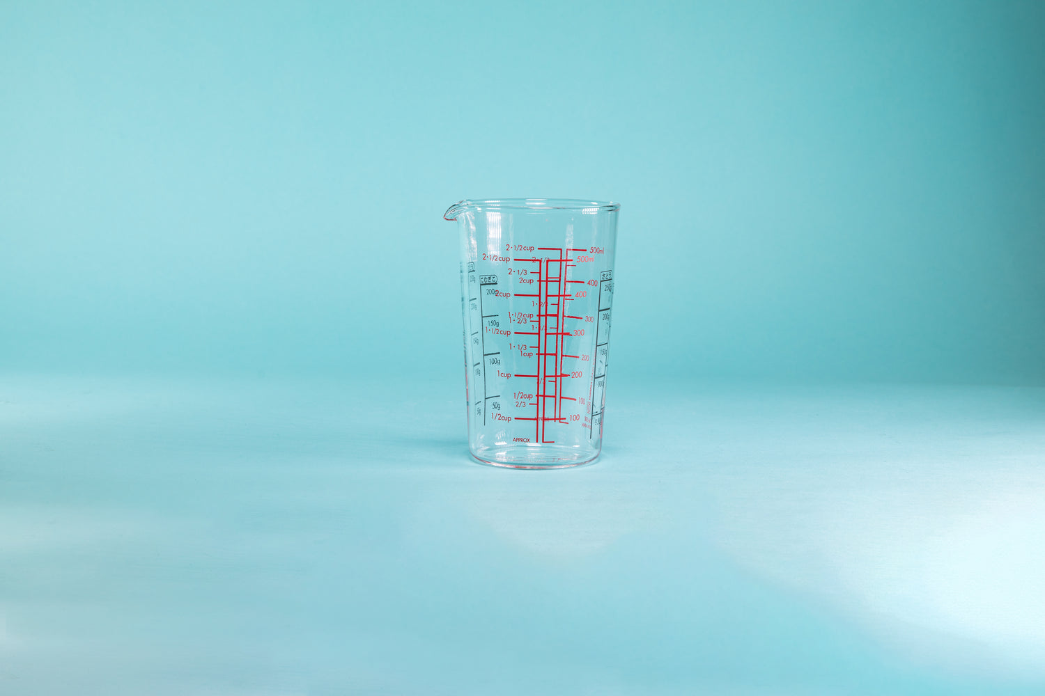 Tall cylindrical measuring cup with small spout and measurement markings in red and black on the inside and outside of the glass set against a blue background.