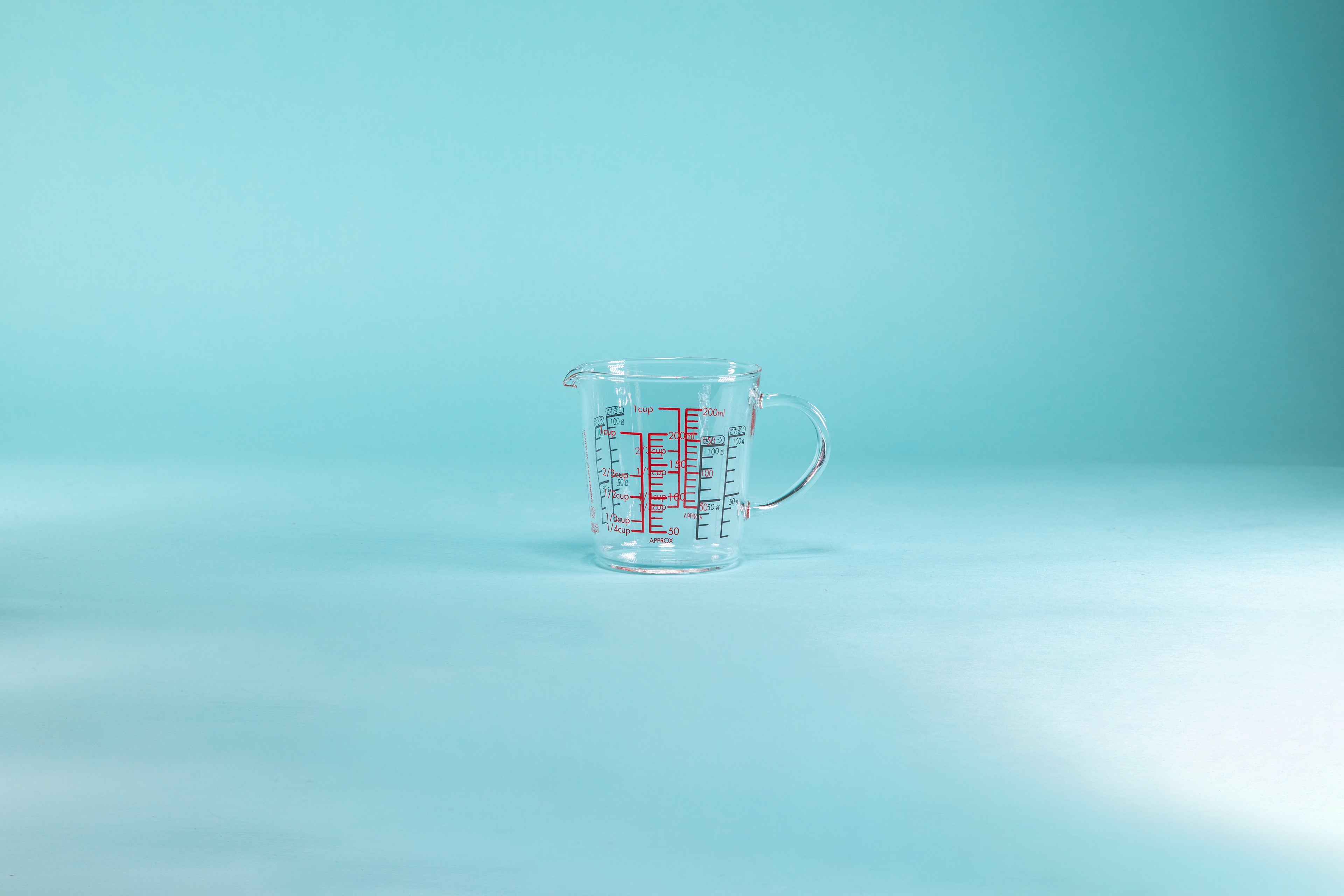 Wide glass measuring cup with spout, full glass handle, and measurements in red and black on the inside and outside of the glass and set against a blue background.