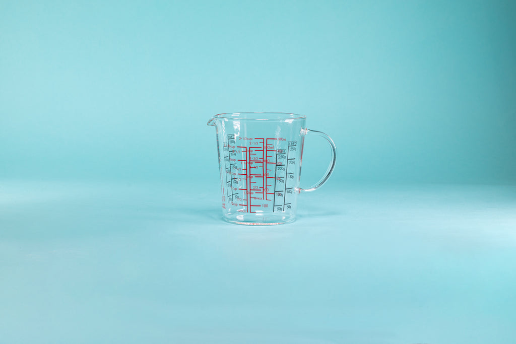 Wide glass measuring cup with spout, full glass handle, and measurements in red and black on the inside and outside of the glass and set against a blue background.