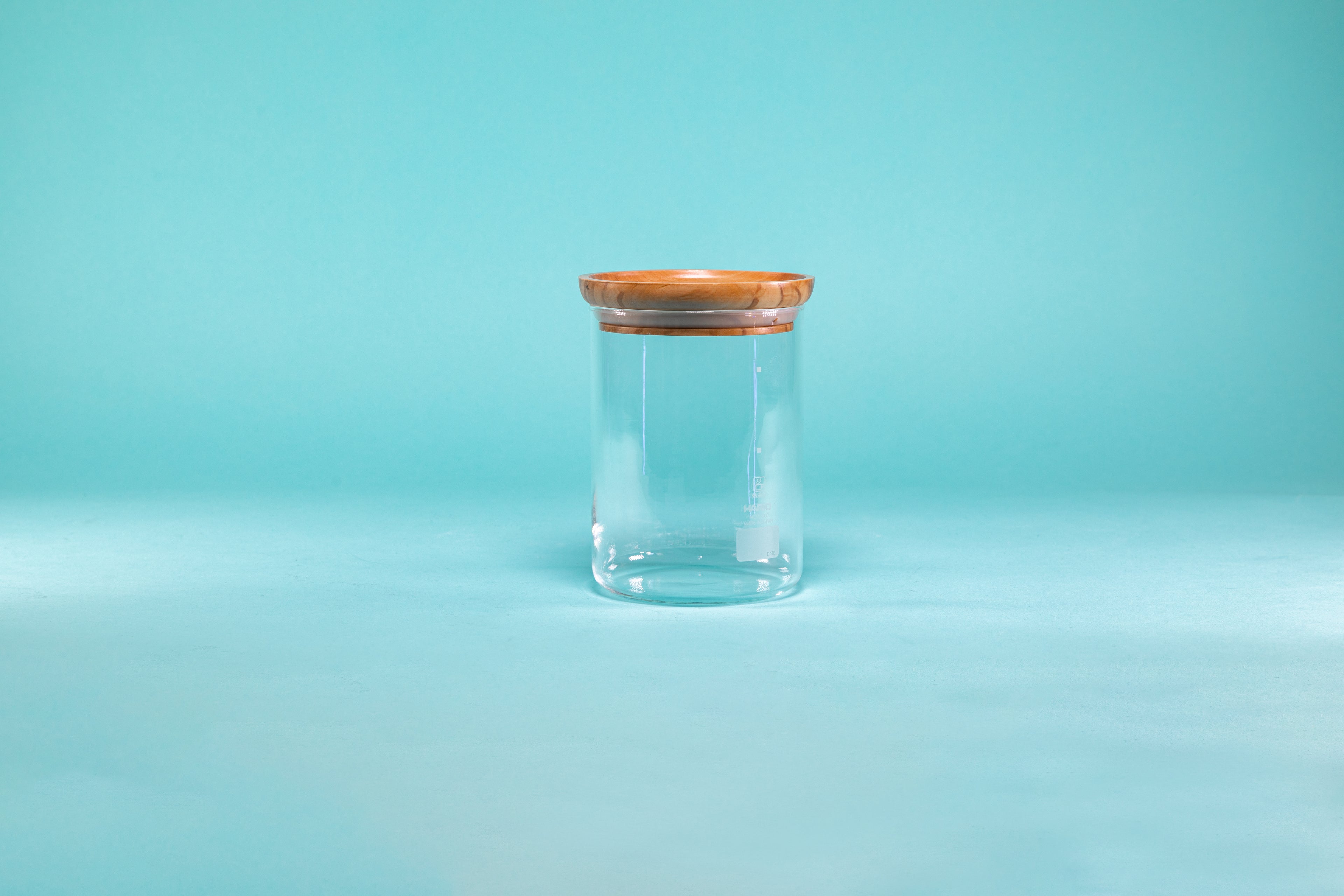 Cylindrical clear glass container with olive wood lid and blue background.