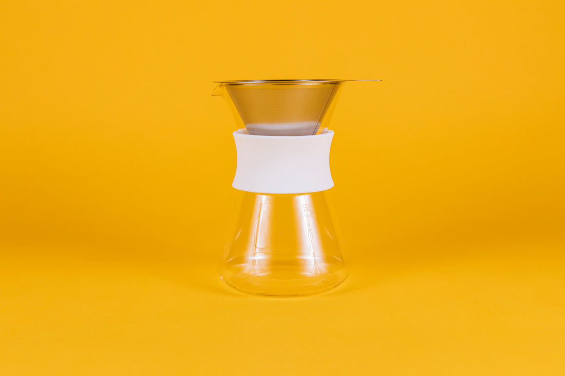 Clear glass server with white silicone collar and stainless steel mesh filter against a yellow background.