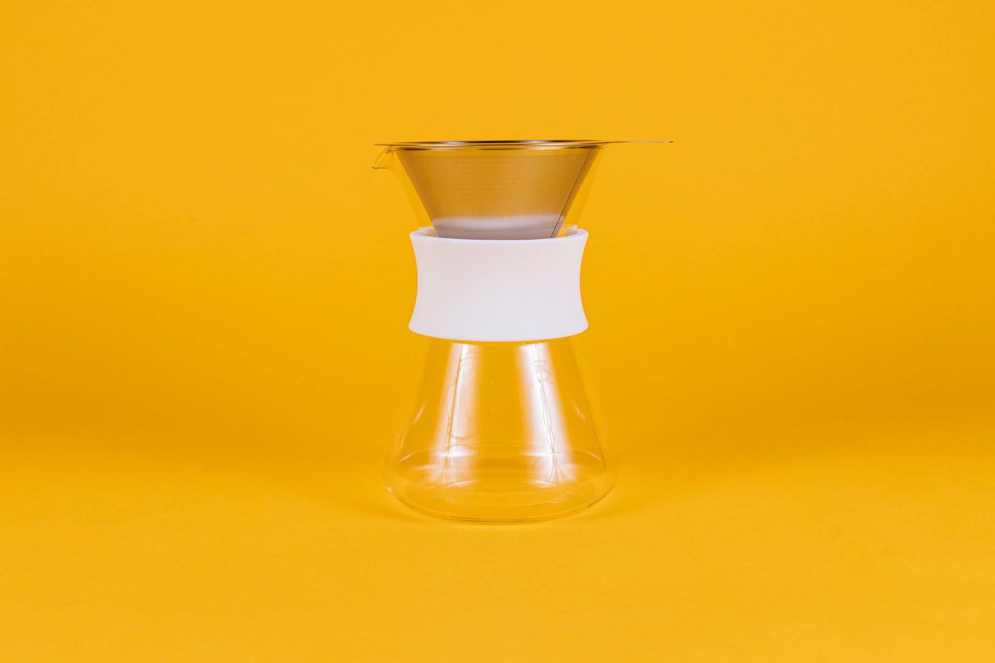 Clear glass server with white silicone collar and stainless steel mesh filter against a yellow background.