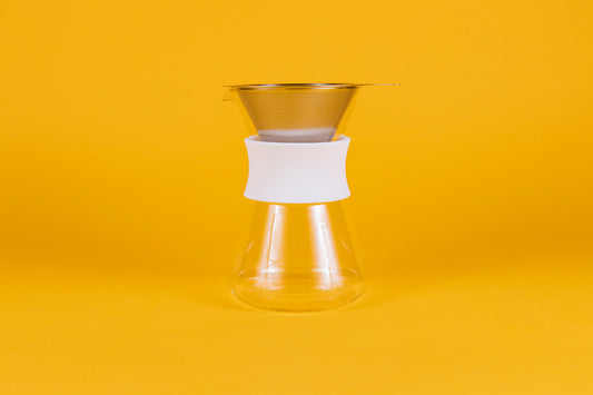 Clear glass server with white silicone collar and stainless steel mesh filter against a yellow background.