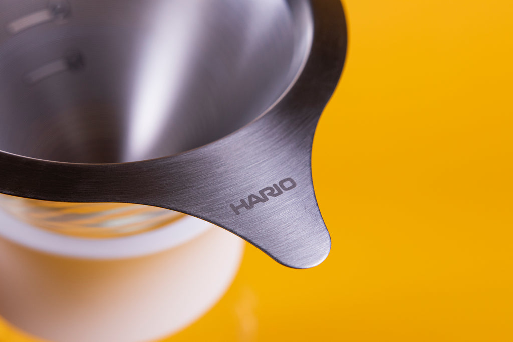 Stainless steel mesh filter with Hario logo on tab and blurred parts of the glass server with white silicone collar against a yellow background.