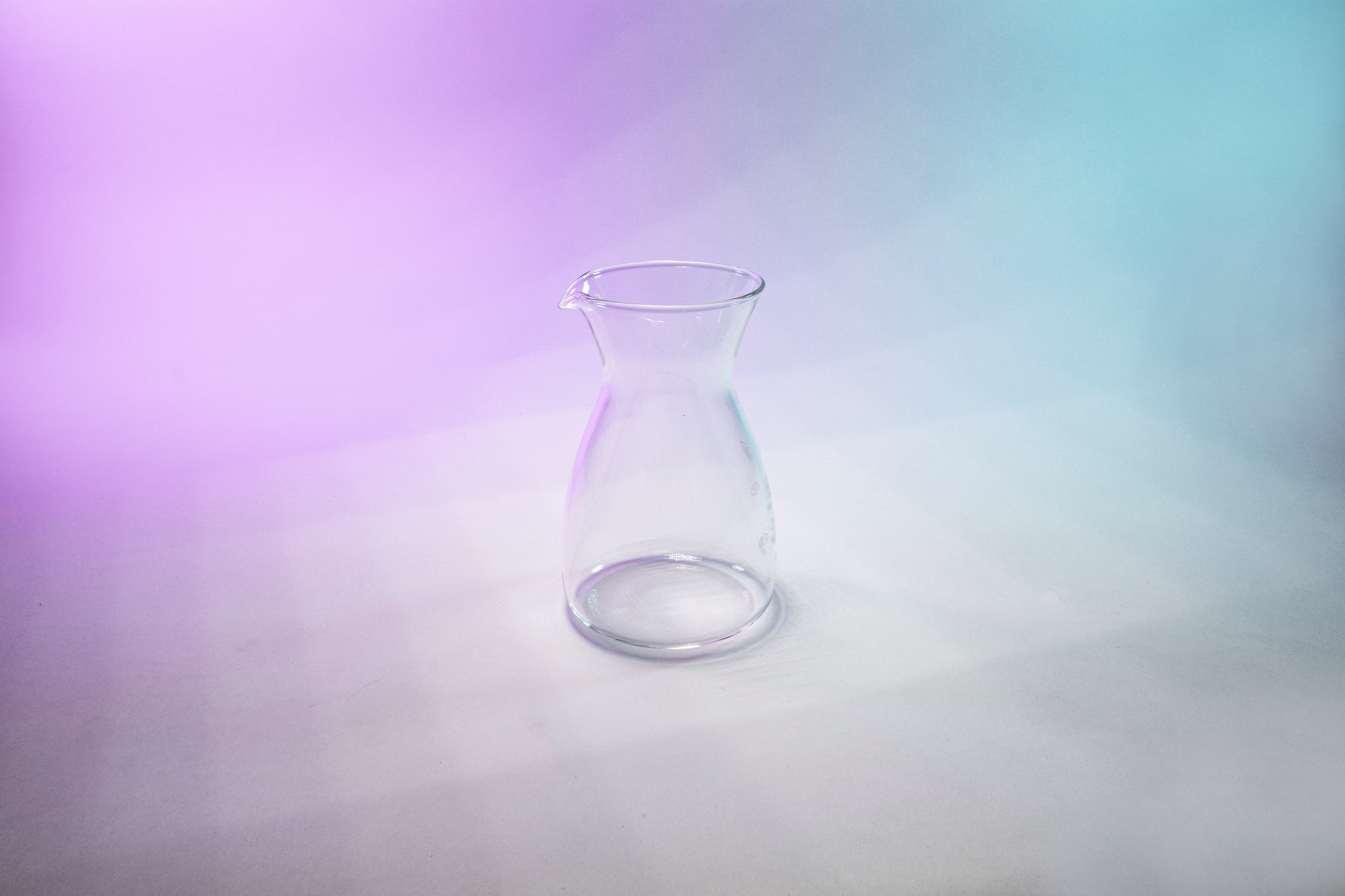 Tapered glass coffee decanter with flared spout against a purple, gey, and blue background.