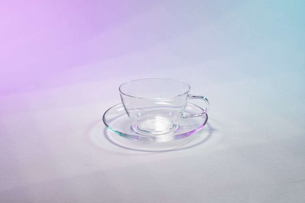 Clear glass tea cup on round glass saucer set against a purple and blue gradient background.
