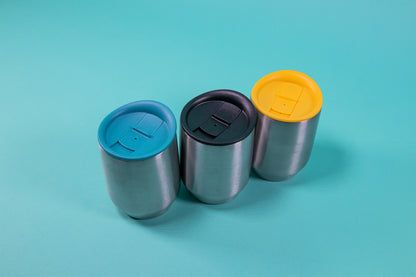 Three short stainless steel mugs with light blue, black, and yellow silicone lids and sliding stoppers set against a blue background.
