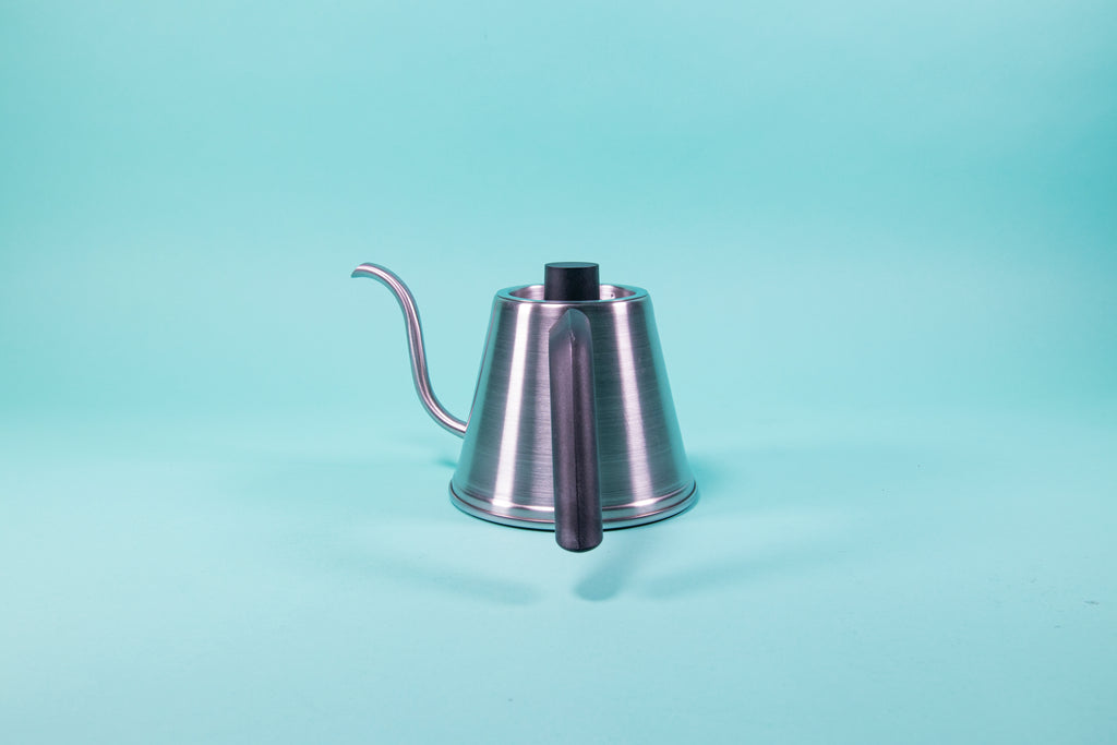 Showing the asymmetric angle of the handle on a silver stainless steel kettle with black plastic handle and short cylindrical black plastic lid knob set against a blue background.