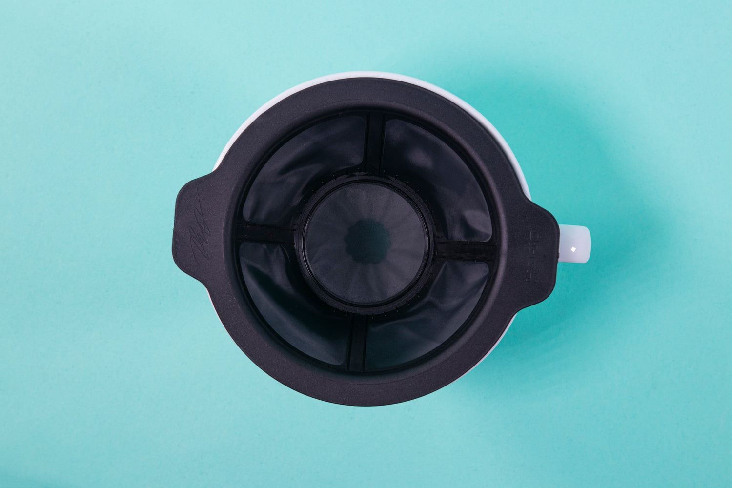 Aerial view of the black plastic filter insert and white ceramic ridges of the white cone shaped dripper on a blue background.