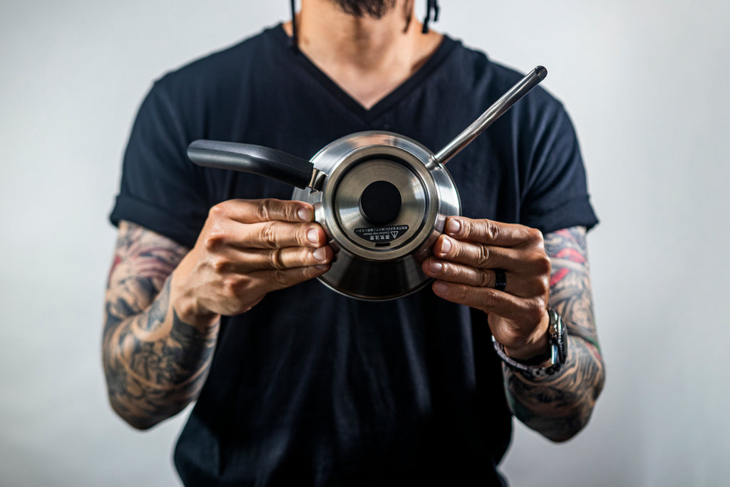 Model in a black t-shirt, with sleeve tattoos, holding a silver stainless steel kettle with black plastic handle and short cylindrical black plastic lid knob set against a light background. Kettle is positioned in the model's hands to show the top of the kettle and the shortened distance between the spout and the handle.