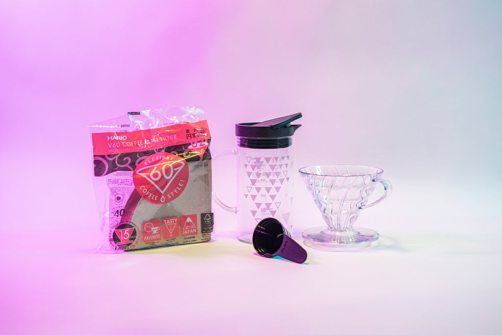 Brown paper cone shaped filters in plastic bag, black plastic measuring spoon, glass server with thermocolor graphics and black plastic lid with cap, and clear plastic cone shaped dripper. Set against a pink gradient background.