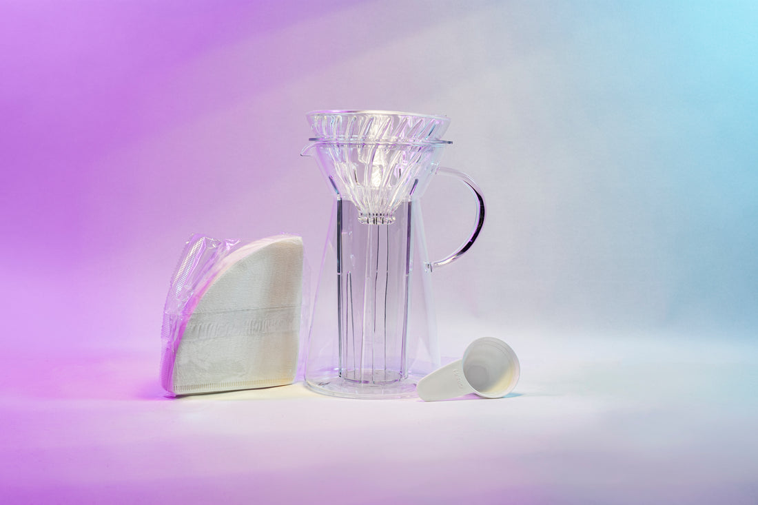 White cone shaped paper filters in a clear plastic bag next to a tapered glass dripper with fluted spout and all glass handle with a cone shaped glass dripper with ribs seated inside, and a white plastic coffee measuring spoon.