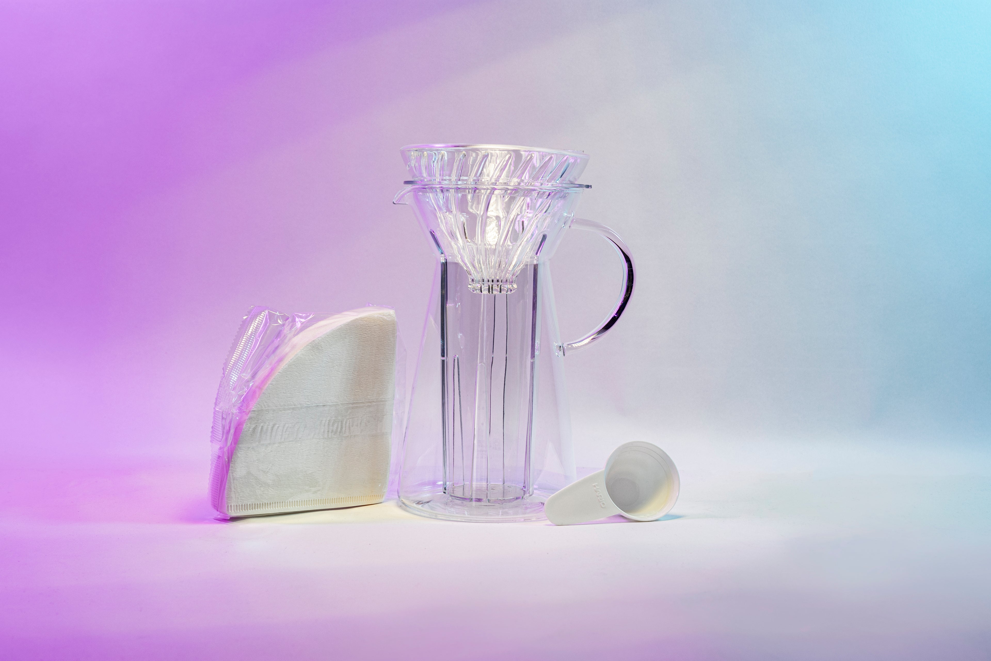 White cone shaped paper filters in a clear plastic bag next to a tapered glass dripper with fluted spout and all glass handle with a cone shaped glass dripper with ribs seated inside, and a white plastic coffee measuring spoon.