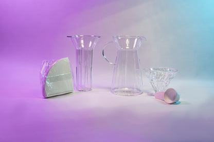 White cone shaped paper filters in a clear plastic bag next to a clear plastic ice insert, tapered glass server with fluted spout and all glass handle, glass cone shaped dripper with clear silicone collar, and white plastic measuring spoon.