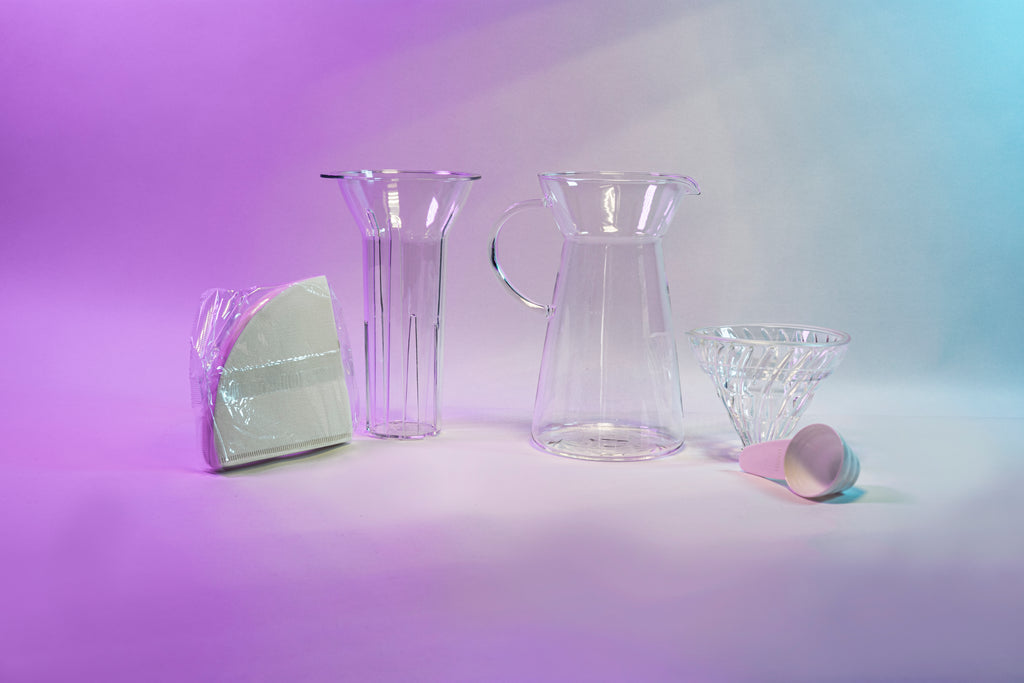 White cone shaped paper filters in a clear plastic bag next to a clear plastic ice insert, tapered glass server with fluted spout and all glass handle, glass cone shaped dripper with clear silicone collar, and white plastic measuring spoon.