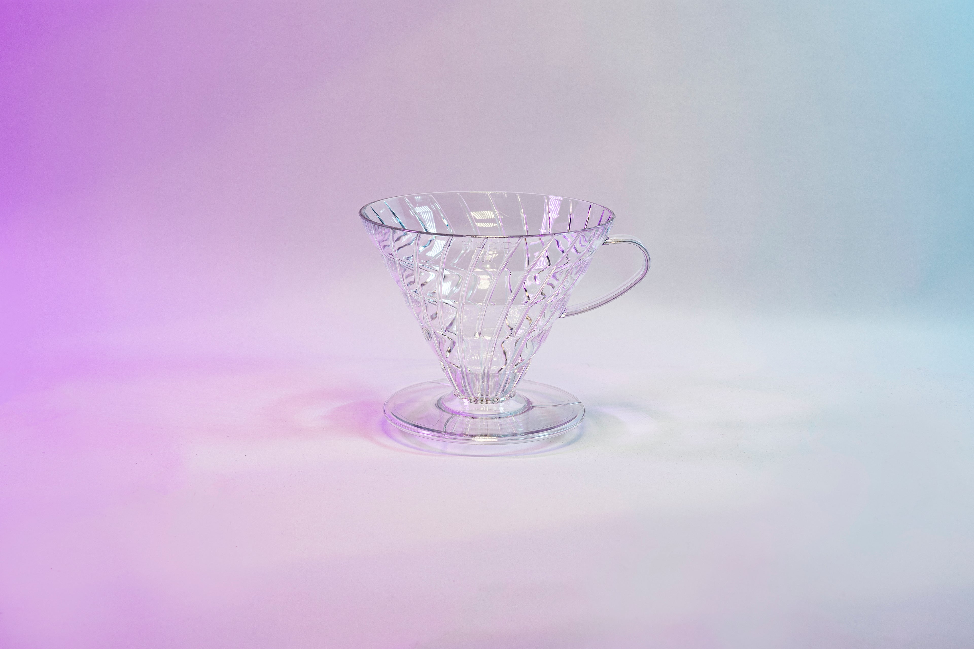 Clear plastic cone shaped dripper with round base and handle on a pink gradient background.