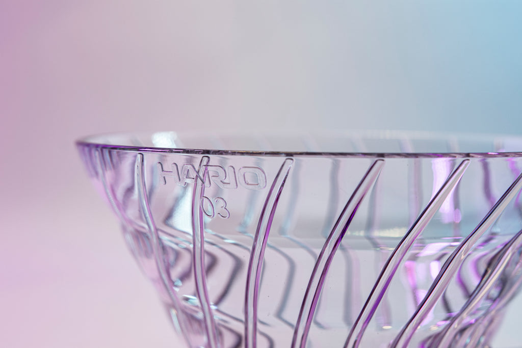 Close up of Hario logo and "03" size number on a clear plastic cone shaped dripper with round base and handle on a pink gradient background.