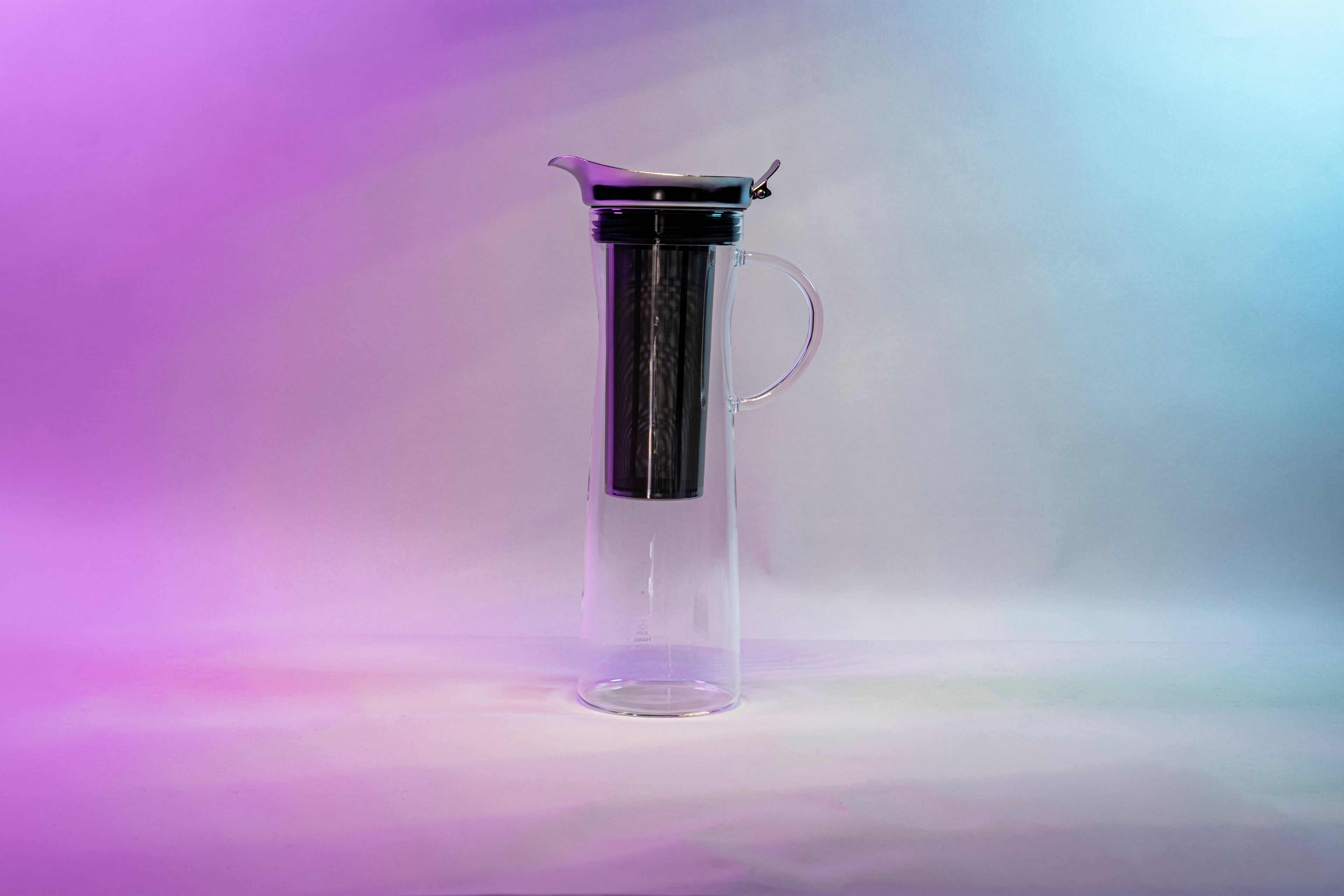 Tall, slim glass coffee pitcher with all glass full handle, stainless steel filter basket, metal lid with flared spout, and black silicone rubber seal against a purple, white, and blue background.
