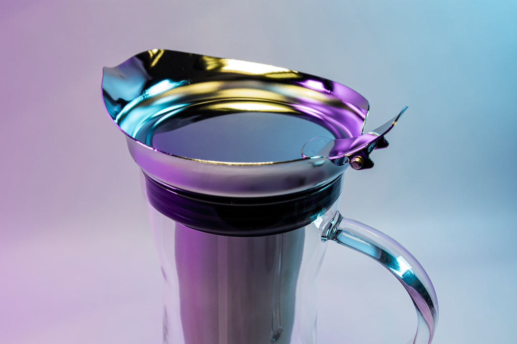 Close up of stainless steel flip-top lid with fared spout on a tall, slim glass coffee pitcher with all glass full handle, stainless steel filter basket, metal lid with flared spout, and black silicone rubber seal against a purple, white, and blue background.