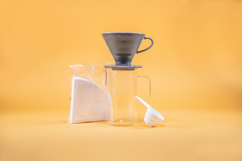 Grey ceramic coffee dripper sitting atop glass coffee server with millileter markings from 100 to 600 and full glass handle, white paper triangular shaped coffee filters in clear plastic bag, and white plastic coffee measuring spoon against a yellow background.